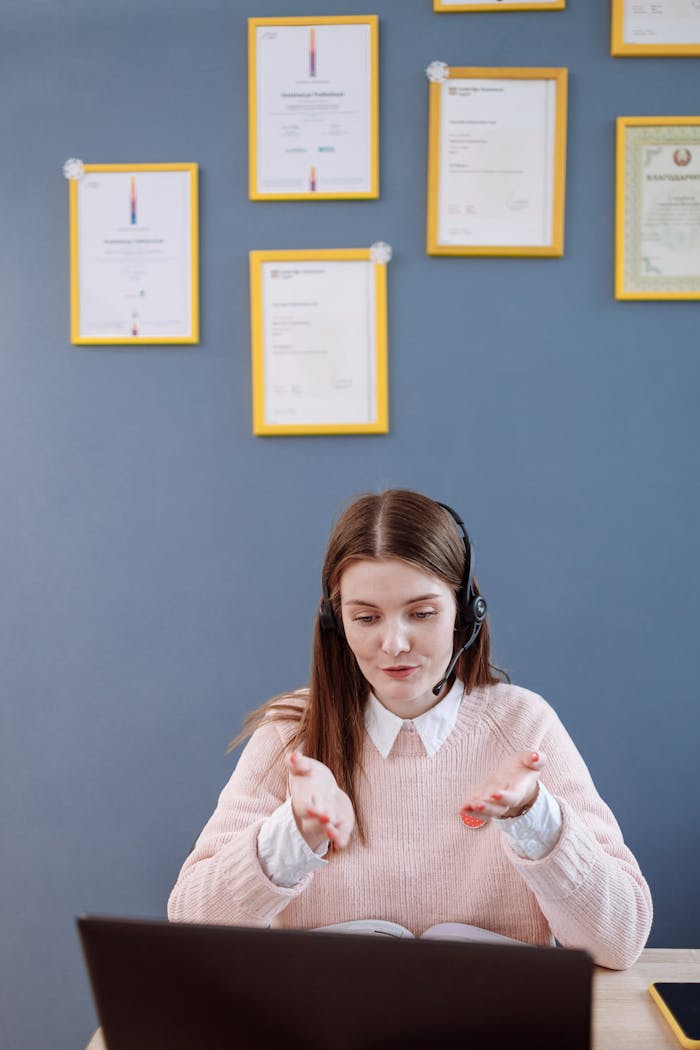 A woman wearing a headset having an online meeting, with certificates framed on the wall behind.